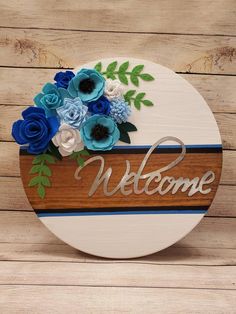 a welcome sign with blue flowers and greenery on it sitting on a wooden surface