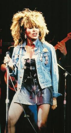 a woman standing in front of microphones on stage wearing a jean jacket and skirt