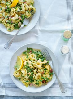 two plates of pasta with peas and lemon wedges on a white table cloth next to silverware