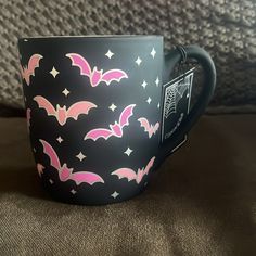 a black mug with pink bats and stars on it sitting on a couch next to a pillow