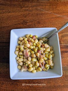 a white bowl filled with beans and ham on top of a wooden table next to a spoon