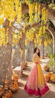 a woman in a yellow and pink dress standing under an archway with flowers hanging from it