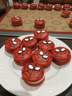 red spiderman cookies are on a white plate next to a tray of cupcakes