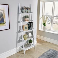 a white leaning shelf with books and plants on it in front of a large window