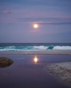 a full moon is seen over the ocean and beach at dusk with waves coming in