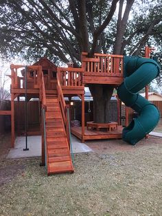 a wooden play set with a green slide next to a large tree in the yard