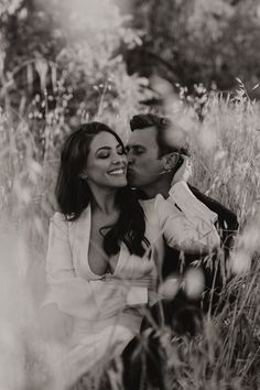 a man and woman sitting in tall grass kissing each other with their heads close to the ground