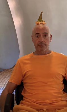 a bald man in an orange shirt with a golden crown on his head sitting in a chair