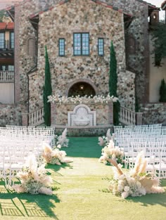 an outdoor ceremony setup with white chairs and flowers on the grass in front of a stone building