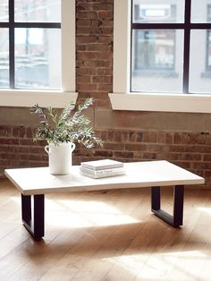 a white coffee table with a plant on it in front of two windows and a brick wall