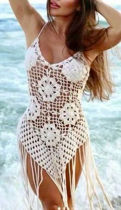 a woman in a white crochet swimsuit standing on the beach with her arms behind her head