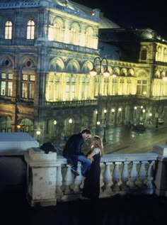 a man and woman are sitting on a balcony overlooking a city street at night with buildings lit up in the background