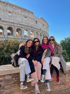 three women sitting on the side of a brick wall in front of an ancient building