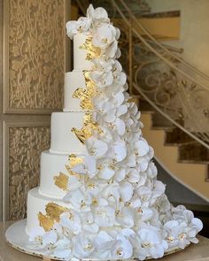 a white and gold wedding cake with flowers on the bottom tier is displayed in front of a staircase