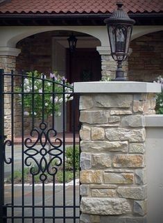 an iron gate with a clock on it in front of a brick building and stone pillars