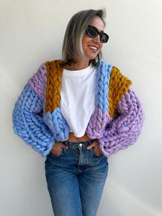 a woman standing against a wall with her hands in her pockets and wearing jeans, a white t - shirt and colorful knitted cardigan