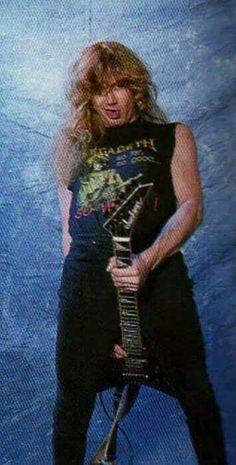 a man with long hair holding a guitar in front of a blue wall and posing for the camera