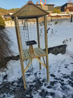 a wooden bird feeder in the snow next to a fence and bushes with no leaves on it