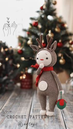 a knitted reindeer doll standing in front of a christmas tree on a wooden table