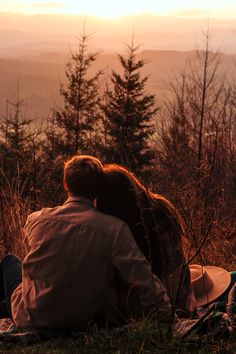 two people are sitting on the ground in front of some trees and mountains at sunset