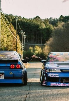 two cars parked next to each other on a street with trees in the background and one car has its hood up