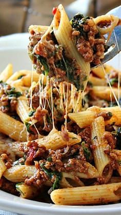 a fork full of pasta with meat and spinach on it, being lifted from a white bowl