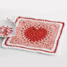 a crocheted square with a red heart on it next to a bowl of sprinkles