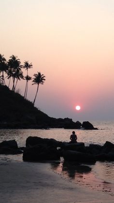 a person sitting on rocks in the water at sunset with palm trees behind them and an orange sky