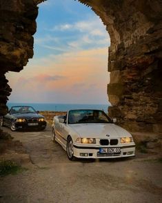 two cars are parked in front of an arch on the road near the ocean at sunset