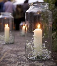 two glass jars filled with white flowers and lit candles sitting on a stone path in front of people