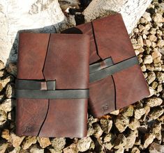 two brown leather notebooks sitting next to each other on top of some rocks and logs