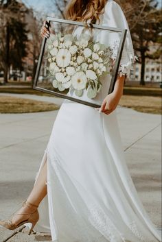 a woman in a white dress is holding a framed flower arrangement with her legs crossed