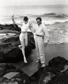 two people standing on rocks near the ocean with their arms in the air and one person holding something up