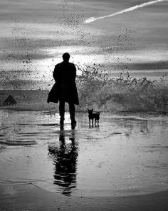 a black and white photo of a man walking his dog on the beach at sunset