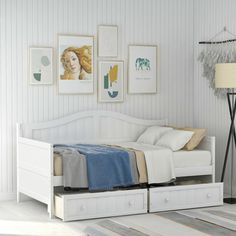 a white bed with drawers underneath it in a room next to a lamp and pictures on the wall