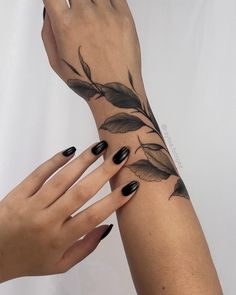 two hands with black nail polish holding onto each other's wrist and one has a leaf tattoo on it