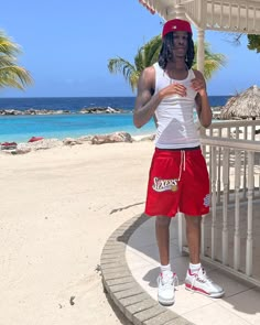 a man in red shorts and a white tank top standing under an umbrella on the beach