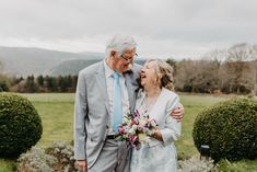 an older man and woman standing next to each other in front of some bushes with flowers