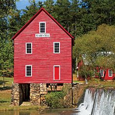 a red building with a waterfall in front of it and trees around the water's edge