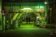 two people are walking under an overpass with graffiti on the walls and green lights