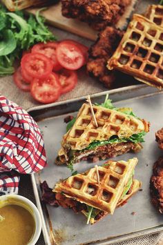 fried chicken and waffle sandwiches on a tray next to a cup of sauce, tomatoes and lettuce