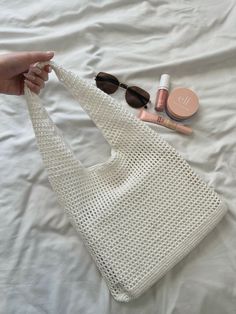 a hand holding a white purse next to some makeup and eyeglasses on top of a bed