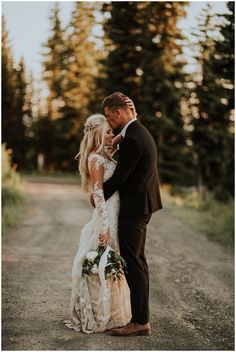 a bride and groom standing in the middle of a dirt road with trees behind them