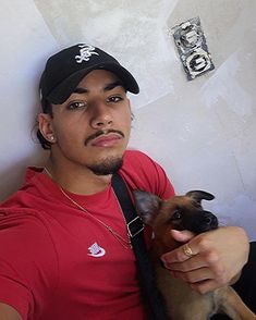 a man in a red shirt holding a small brown and black dog while wearing a baseball cap