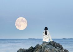 a woman sitting on top of a rock next to the ocean under a full moon