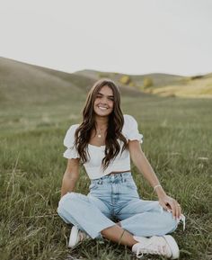 a woman sitting on the ground in a field smiling at the camera with her legs crossed