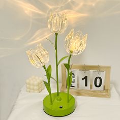 a green clock and three flowers on a white tablecloth with lights in the background