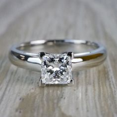 a white gold ring with a princess cut diamond in the center on a wooden surface