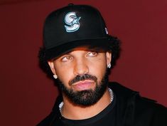 a man with a beard wearing a black baseball cap and looking at the camera while standing in front of a red wall