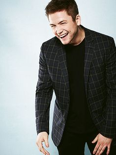 a man laughing while standing in front of a blue background wearing a black t - shirt and plaid blazer